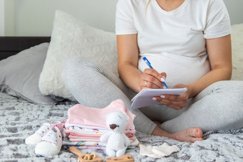 Pregnant woman writing newborn hospital bag checklist with baby items around.