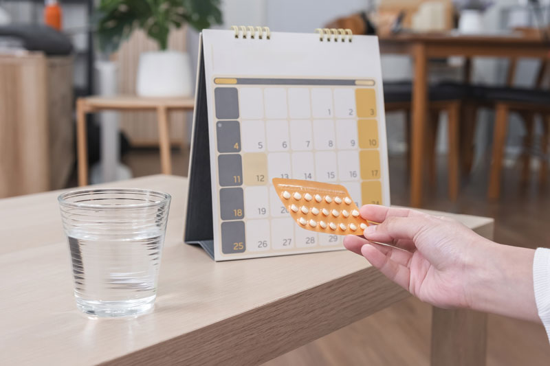 Hand holding prenatal vitamins near a calendar and glass of water on table
