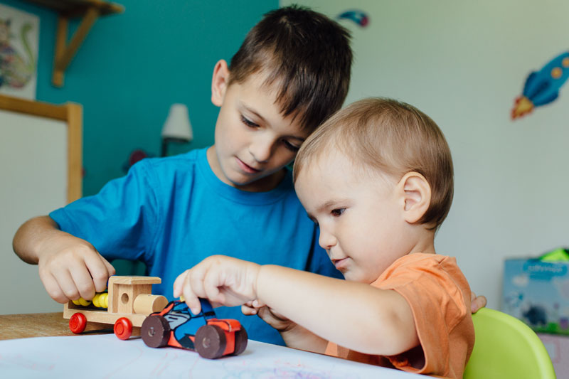 Older brother and younger sibling playing together with wooden toy cars at home