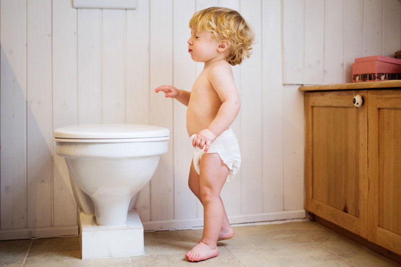 Toddler in diaper standing curiously in front of a white toilet, learning about potty training